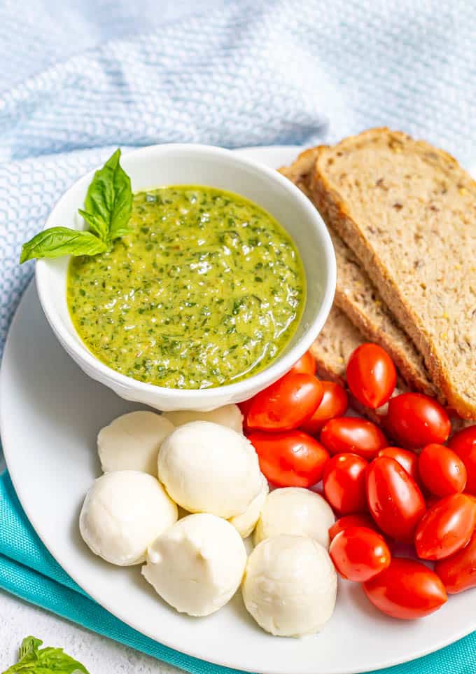 Pesto served in a white bowl on a plate of whole grain bread, tomatoes and mozzarella balls