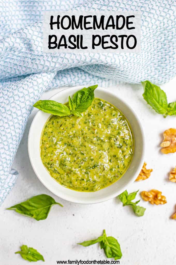 A white bowl of fresh basil pesto with sprigs of fresh pesto and walnut pieces scattered nearby and a text box on top
