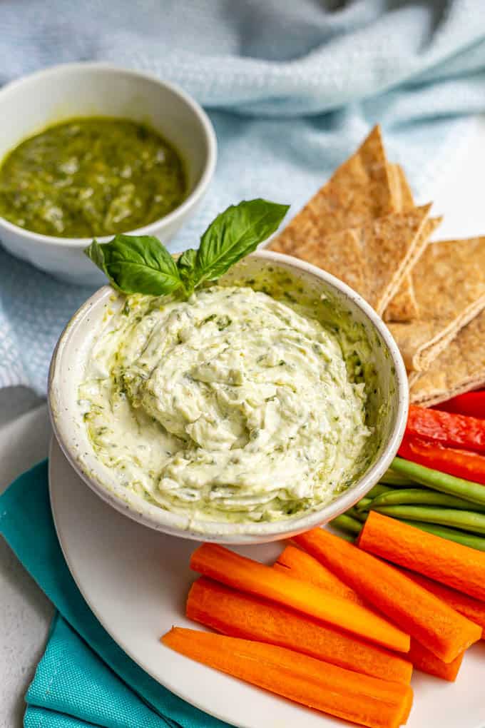 A bowl of pesto Greek yogurt dip with a sprig of fresh basil on a plate of fresh veggie sticks and pita triangles