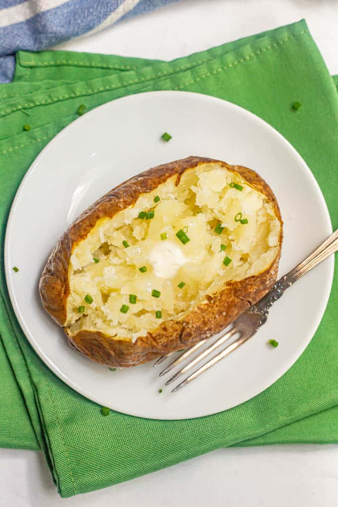A fluffy baked potato with a crispy skin on a white plate with a fork alongside