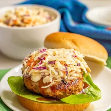 A coleslaw topped burger with BBQ sauce and lettuce and a bowl of coleslaw in the background