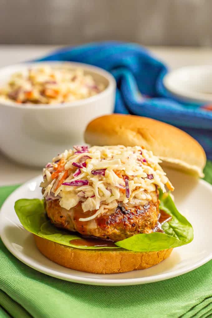 A coleslaw topped burger with BBQ sauce and lettuce and a bowl of coleslaw in the background