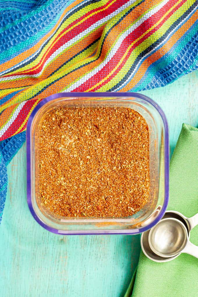 A square glass container filled with a homemade seasoning mix with measuring spoons resting nearby