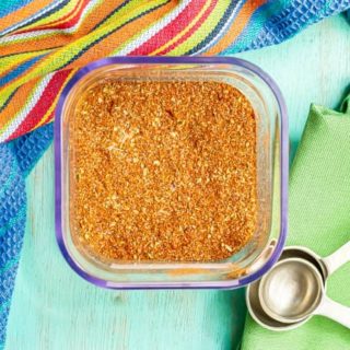 A square glass container filled with a homemade seasoning mix with measuring spoons resting nearby