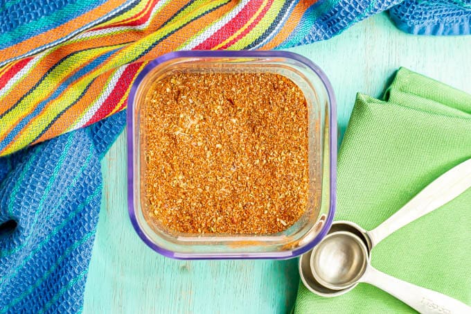 A square glass container filled with a homemade seasoning mix with measuring spoons resting nearby