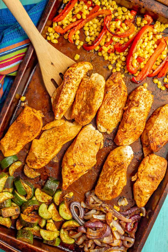 A sheet pan of chicken and vegetables with a wooden spoon lifting up some of the seasoned chicken strips