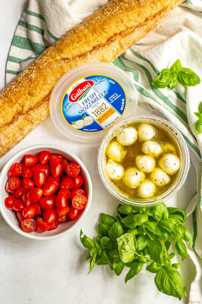 Ingredients laid out on a counter for making bruschetta, including a baguette, fresh tomatoes, marinated mozzarella and fresh basil