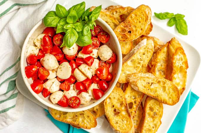 A serving platter of baguette slices with a bowl of marinated mozzarella with tomatoes and basil