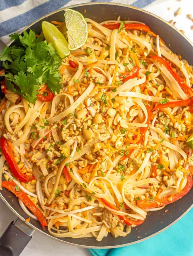 Chicken pad Thai with rice noodles in a large skillet topped with peanuts, cilantro and lime wedges