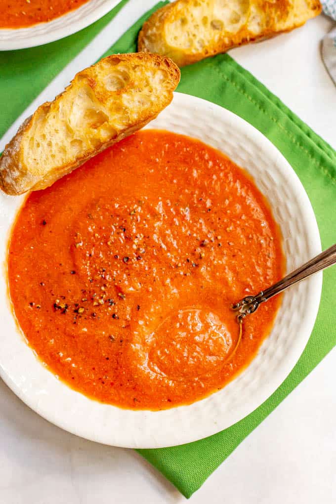 A spoon resting in a bowl of creamy tomato soup with a toasted slice of bread resting on the side of the bowl