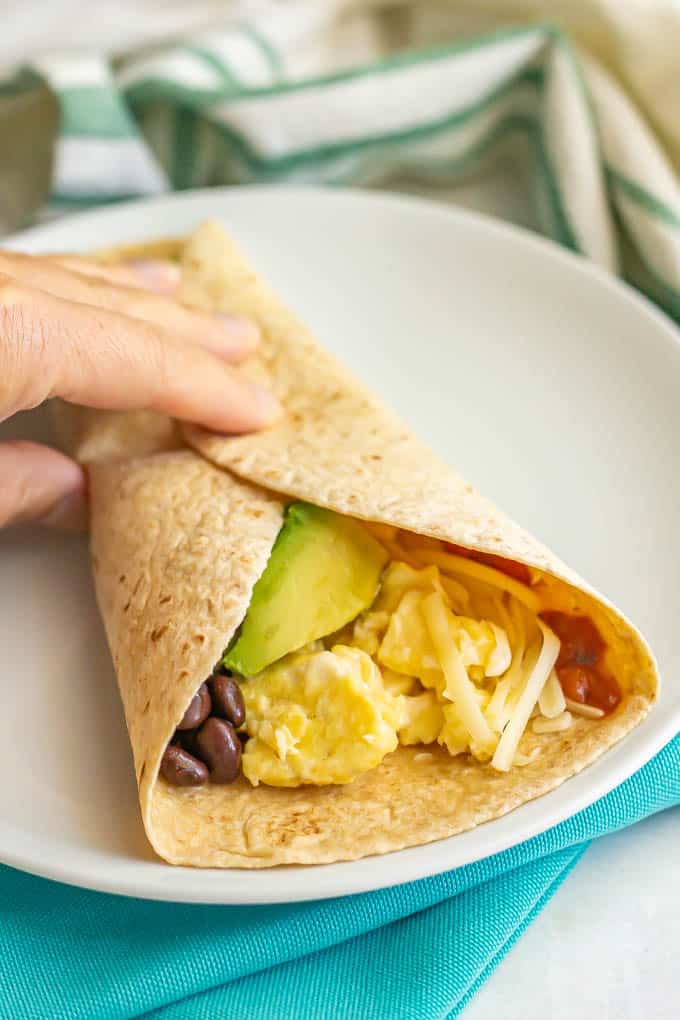 A hand holding a wrapped tortilla filled with scrambled eggs, beans, cheese, salsa and avocado