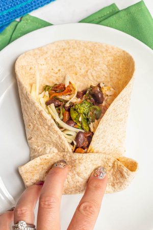 A hand in the process of wrapping up a bean and rice burrito on a white plate
