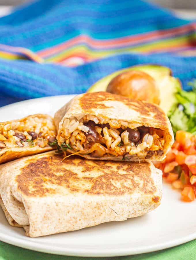 Stacked vegetarian burritos on a white dinner plate with toppings nearby