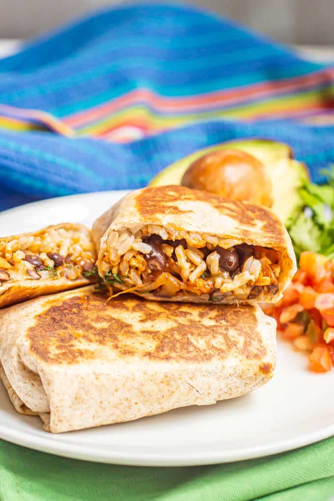 Stacked vegetarian burritos on a white dinner plate with toppings nearby