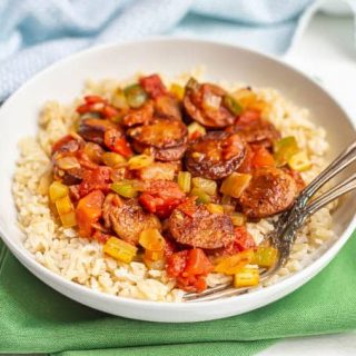 Cajun sausages and veggies served over rice in a large white bowl set on green napkins with forks tucked into the bowl