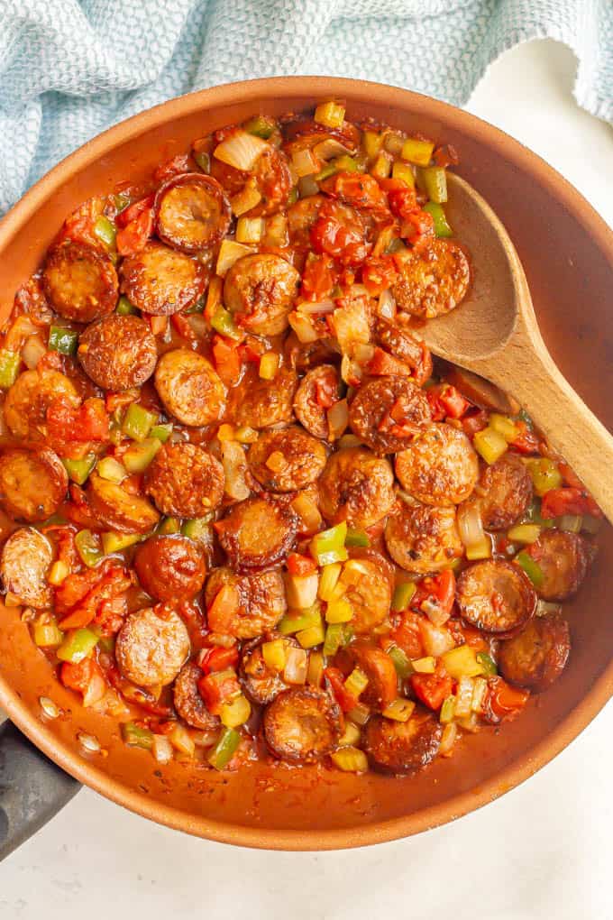 A large skillet with sliced sausages and veggies and a wooden spoon