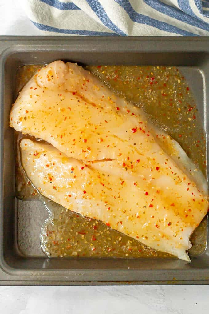 A piece of raw cod in a baking dish covered in Italian dressing before being cooked