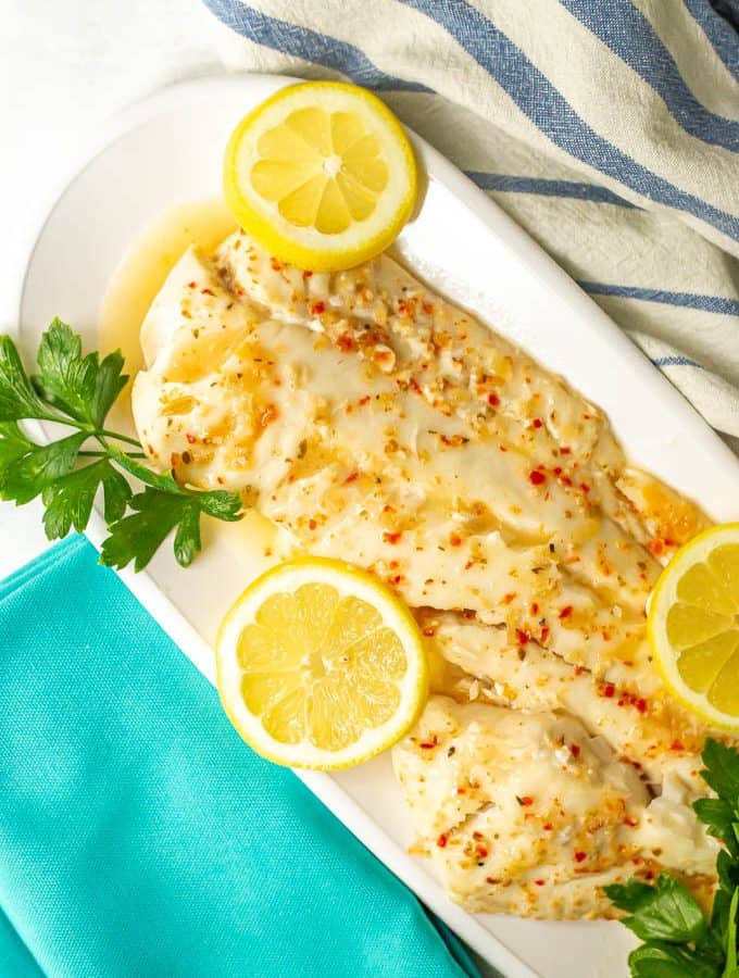 Large fish filet served with sauce on a white serving tray with lemon slices and fresh parsley as garnish