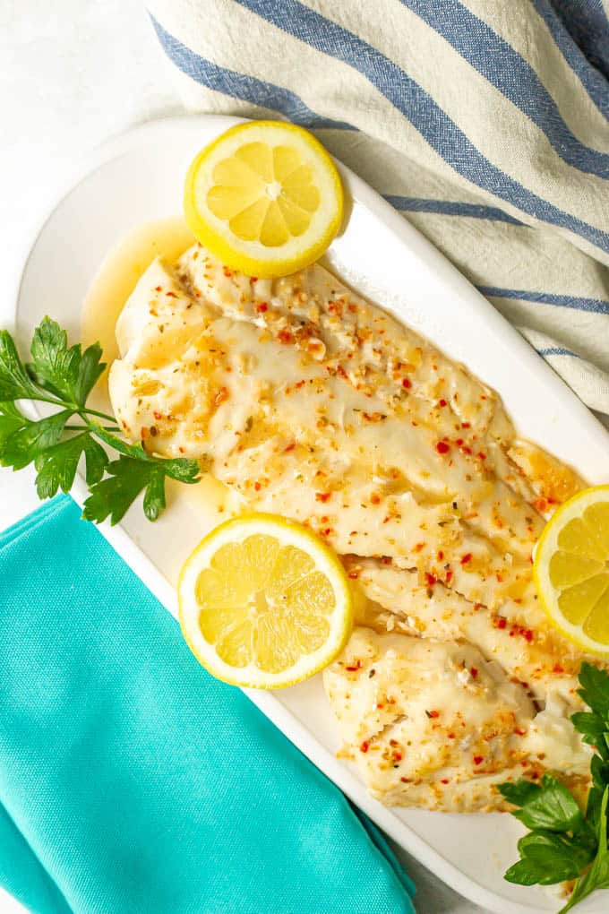 Large fish filet served with sauce on a white serving tray with lemon slices and fresh parsley as garnish