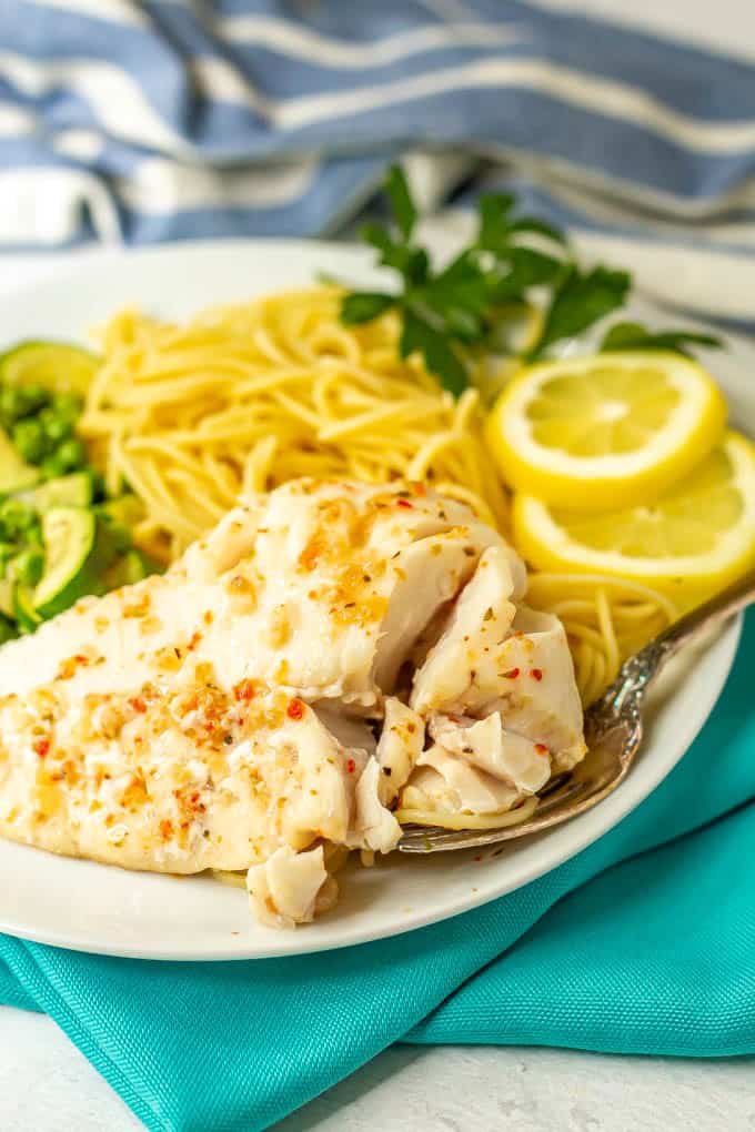 A fork taking a bite of flaky cod with sauce alongside noodles and veggies on a white plate