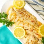 Large fish filet served with sauce on a white serving tray with lemon slices and fresh parsley as garnish and a text box on top of the photo