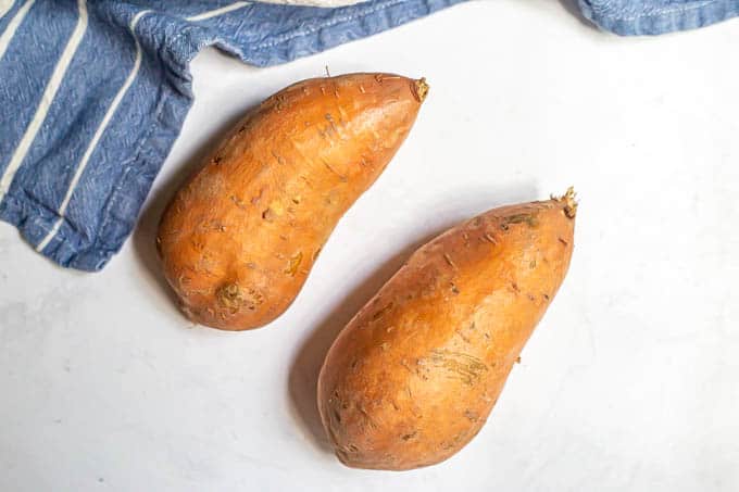 Two scrubbed sweet potatoes on a counter with a blue striped towel nearby