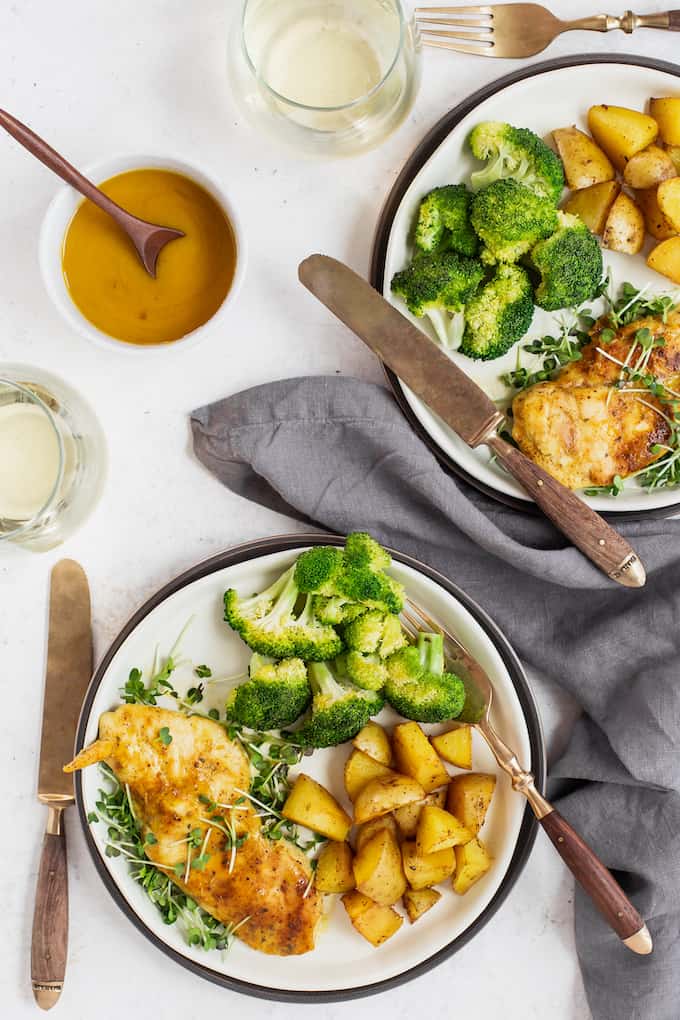 Two plates with honey mustard chicken, broccoli and roasted potatoes, plus utensils and wine glasses