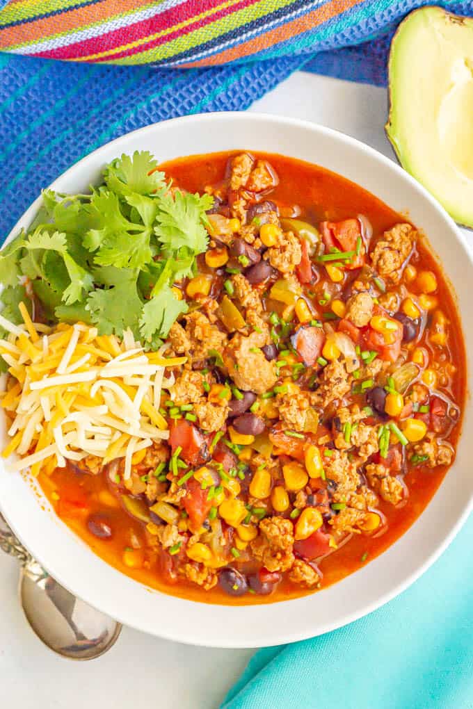 A white serving bowl filled with turkey chili and topped with shredded cheese and cilantro with an avocado half nearby