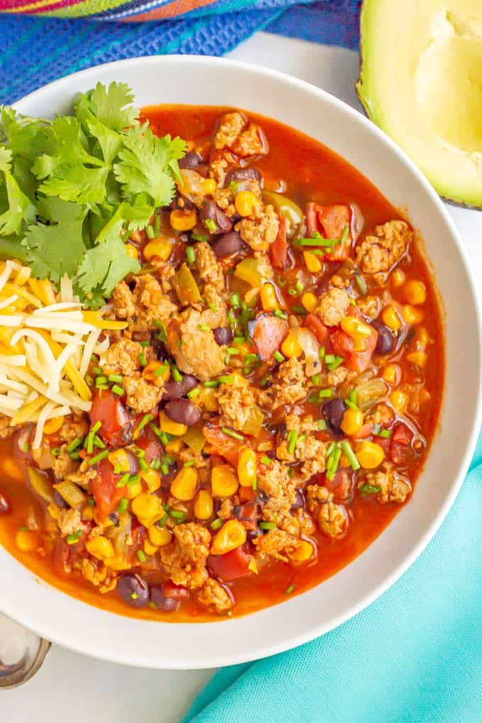 Close up of a colorful ground turkey chili in a white bowl with toppings to the side