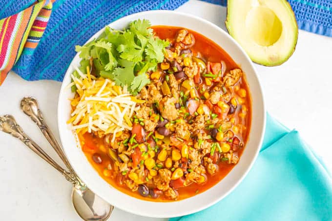 A white bowl of healthy turkey chili with cheese and cilantro and an avocado half, plus spoons nearby