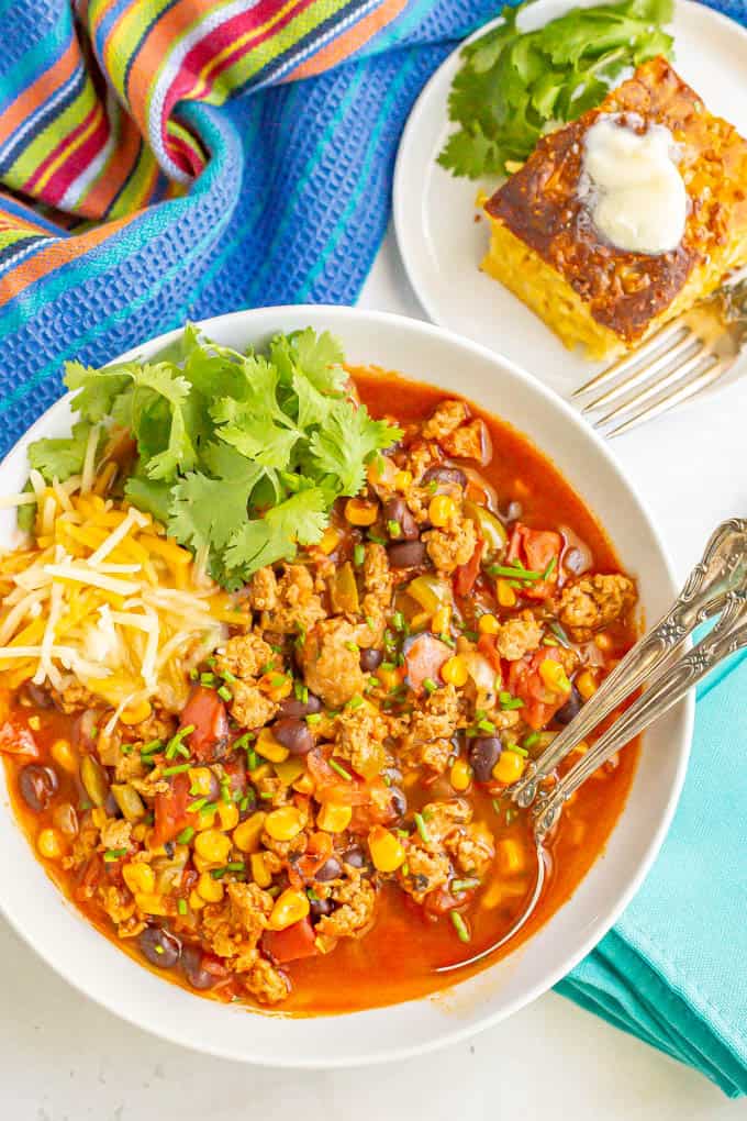 A bowl of turkey chili served with cheese and cilantro and two spoons resting in the bowl with a plate of cornbread nearby