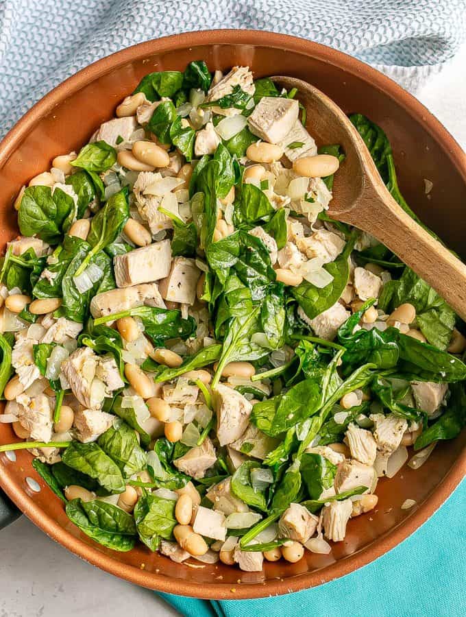 A wooden spoon resting in a copper skillet with a mixture of turkey, white beans and spinach
