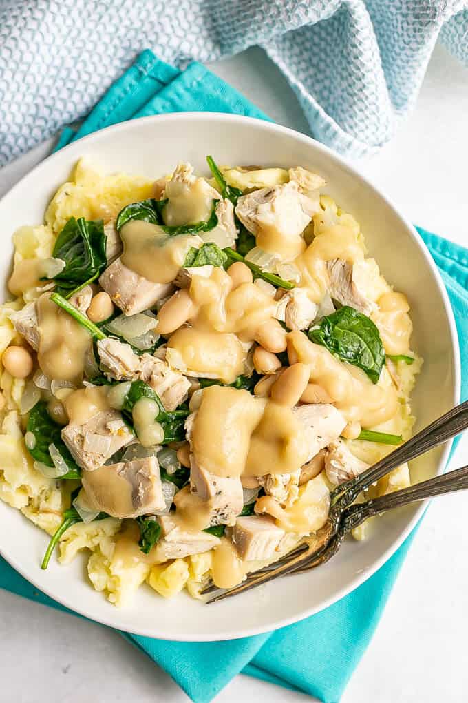 A bowl of mashed potatoes topped with a turkey white bean and spinach mixture with gravy on top and two forks resting in it