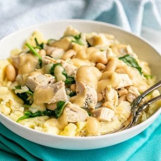 A white bowl of mashed potatoes topped with a turkey white bean and spinach mixture with gravy on top