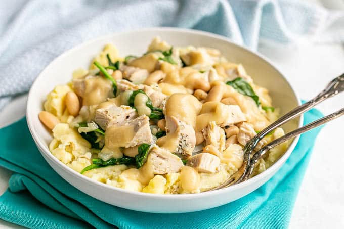 A white bowl of mashed potatoes topped with a turkey white bean and spinach mixture with gravy on top