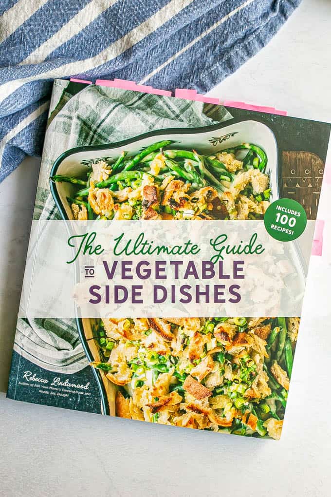 The cover of The Ultimate Guide to Vegetable Side Dishes cookbook on a marble counter with a blue striped dish towel alongside