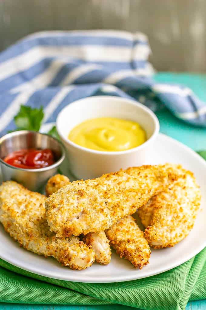 A stack of crispy chicken tenders on a white plate with small bowls of ketchup and honey mustard for dipping