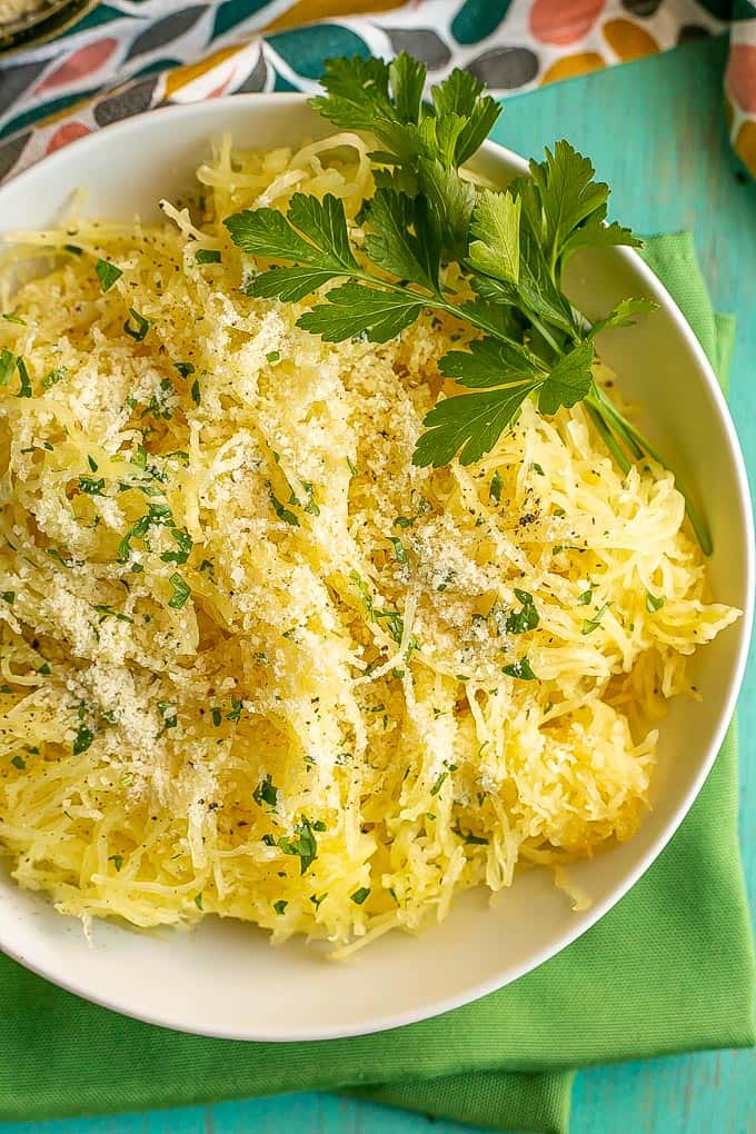 A white bowl full of cooked, pulled spaghetti squash strands sprinkled with Parmesan cheese and parsley with a sprig of parsley on the side