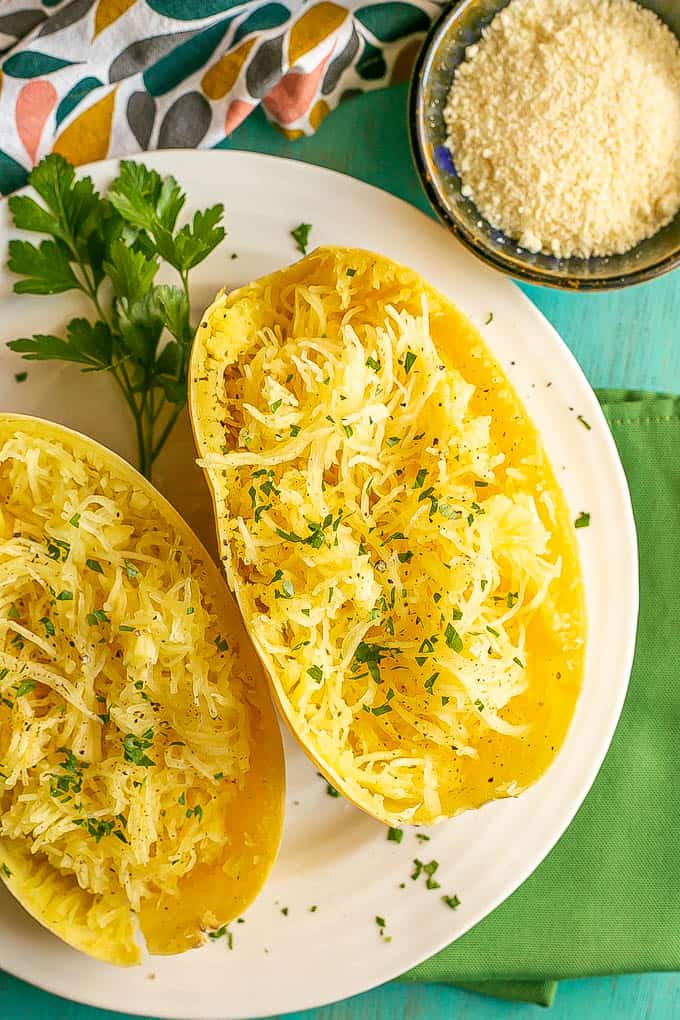 A halved and pulled cooked spaghetti squash sprinkled with parsley and a bowl of Parmesan cheese nearby