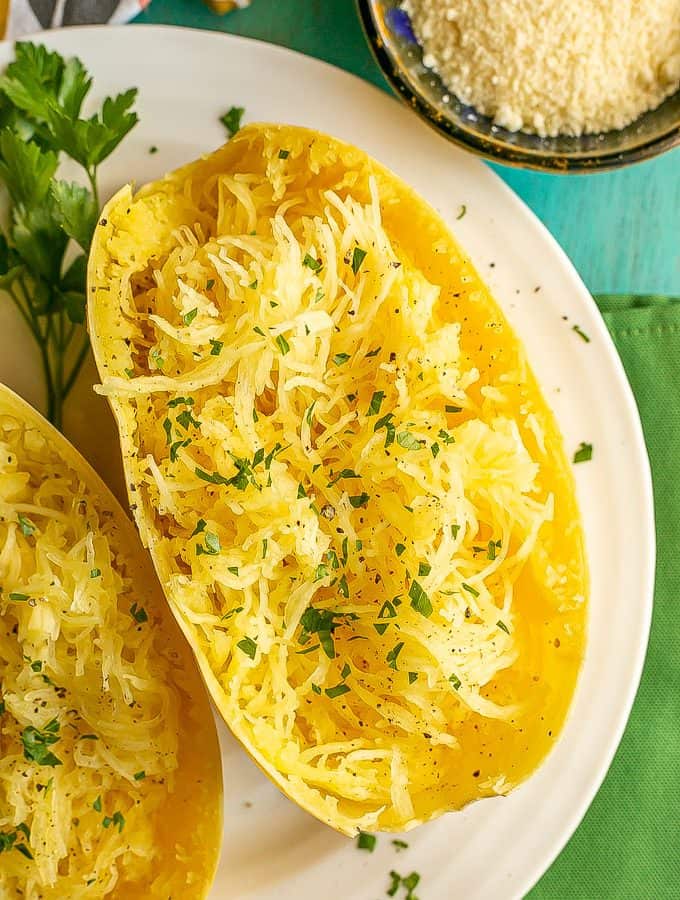 A halved and pulled cooked spaghetti squash with parsley and a bowl of Parmesan cheese nearby