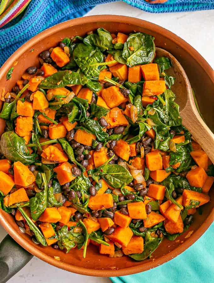 A spoon resting in a skillet of sweet potatoes with black beans and spinach