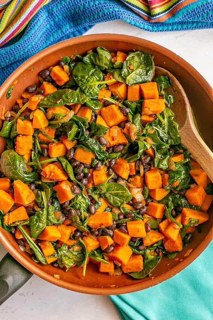 A spoon resting in a skillet of sweet potatoes with black beans and spinach