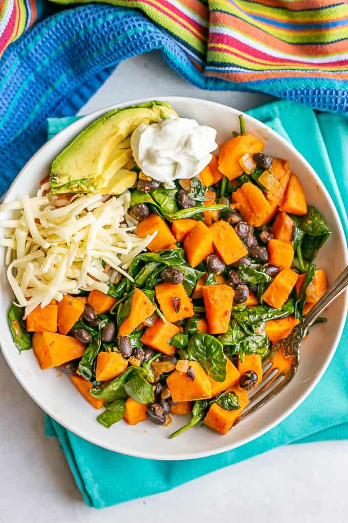 Overhead of a white serving bowl with sweet potatoes, black beans and spinach served with cheese, avocado and Greek yogurt with a fork alongside