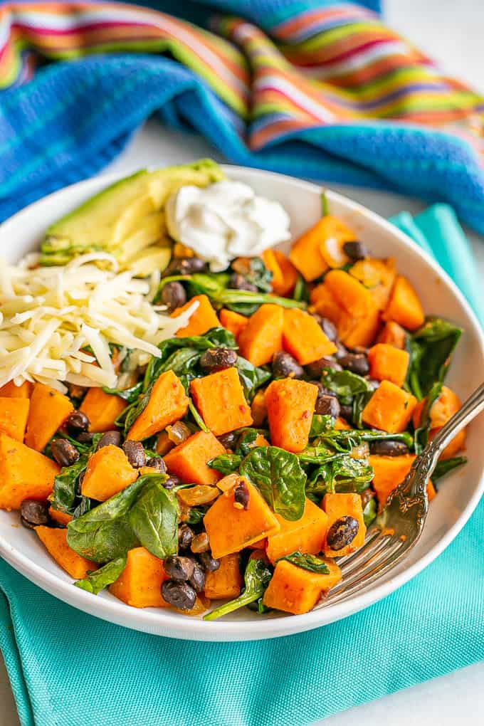 A white serving bowl with sweet potatoes, black beans and spinach served with cheese, avocado and Greek yogurt with a fork alongside