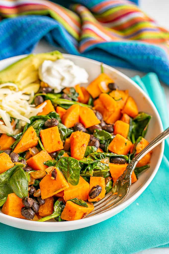 Close up of a vegetarian bowl with sweet potatoes, black beans and spinach, with toppings to the side of the bowl