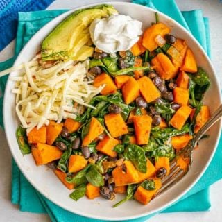 A white bowl with a sweet potato and black bean mixture with wilted spinach, served with shredded cheese, sliced avocado and a dollop of Greek yogurt