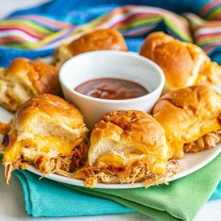 A white plate of cheesy BBQ chicken sliders with a bowl of BBQ sauce in the middle