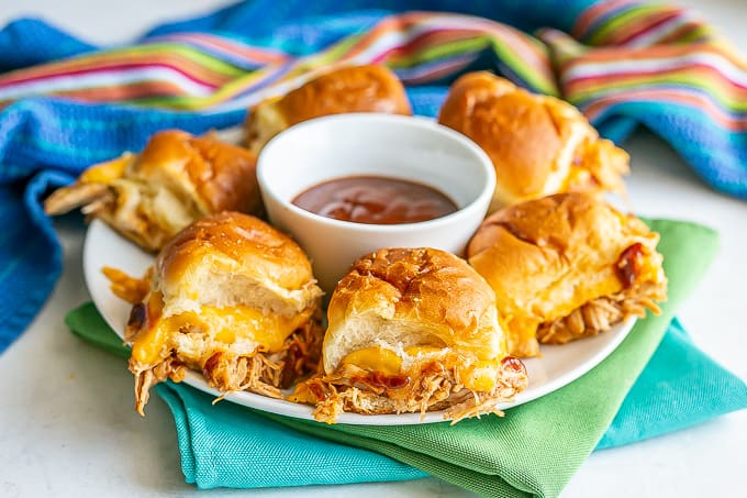 A white plate of cheesy BBQ chicken sliders with a bowl of BBQ sauce in the middle