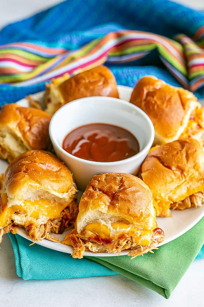 Baked BBQ chicken sliders with melty cheese served on a white plate with a bowl of BBQ sauce for dipping