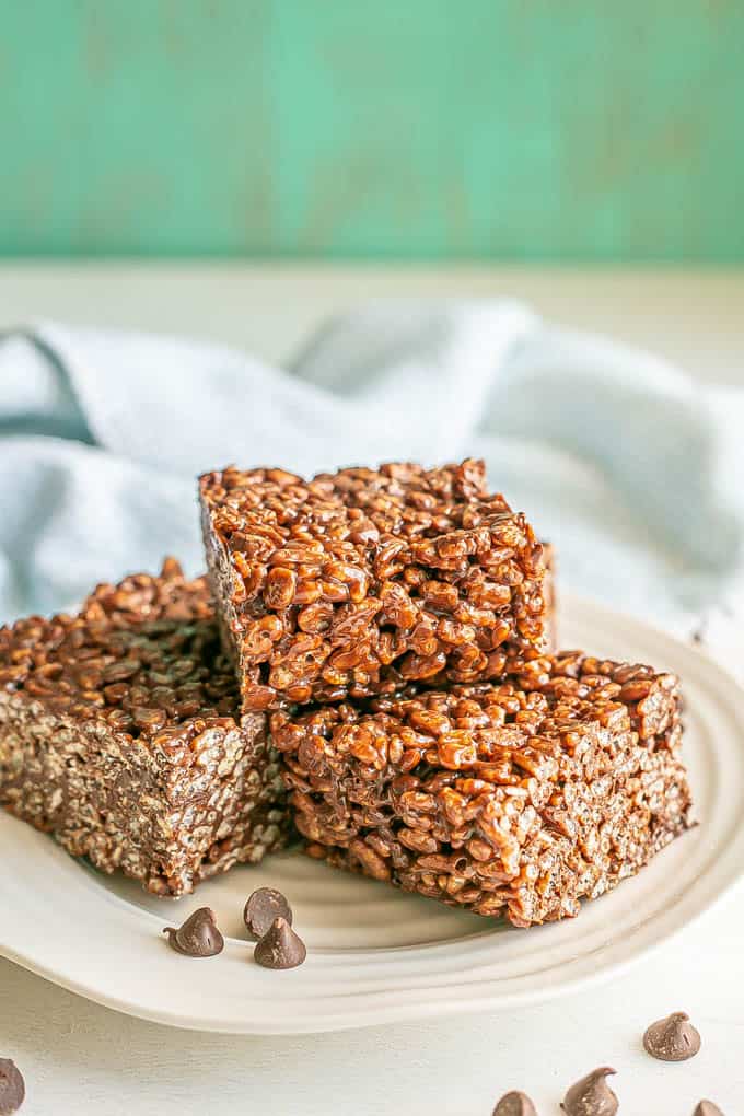 A stack of chocolate rice krispy treats on a white plate with chocolate chips scattered nearby